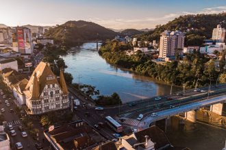 Vista aérea de Blumenau ao entardecer, com edificações, áreas urbanas e o rio cortando a cidade.