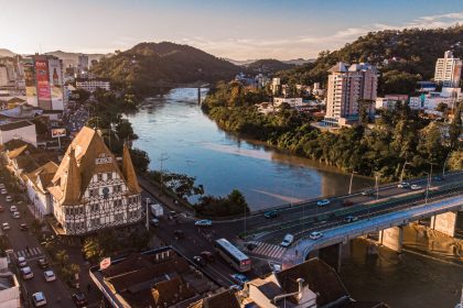Vista aérea de Blumenau ao entardecer, com edificações, áreas urbanas e o rio cortando a cidade.