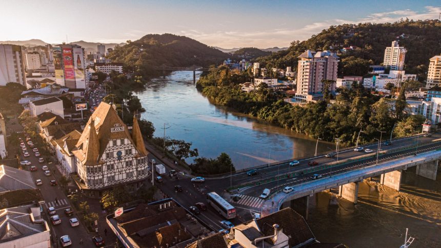 Vista aérea de Blumenau ao entardecer, com edificações, áreas urbanas e o rio cortando a cidade.