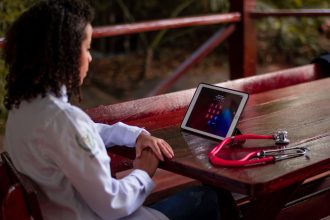 Profissional de saúde usando um tablet e segurando um estetoscópio em uma área externa.