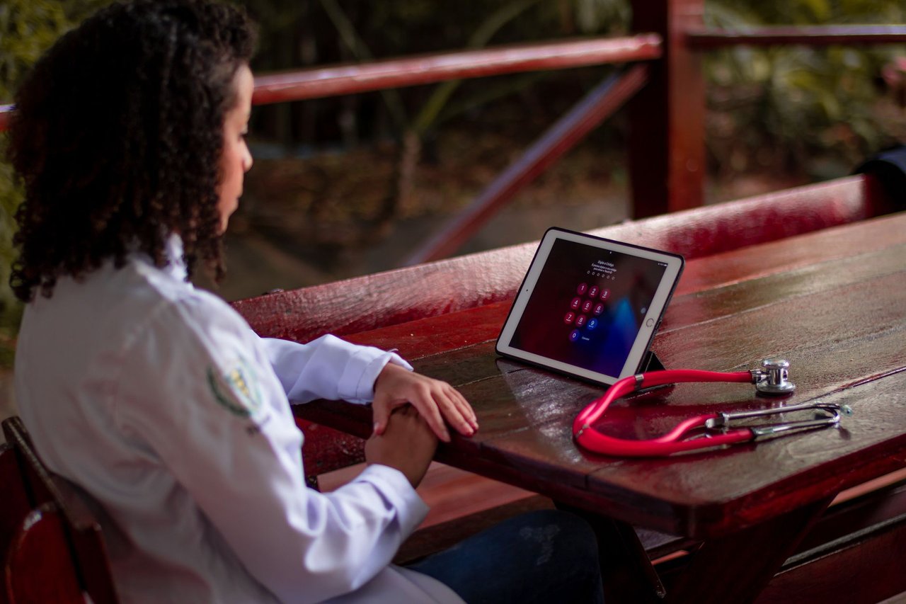 Profissional de saúde usando um tablet e segurando um estetoscópio em uma área externa.