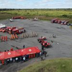 Vista aérea de treinamento de bombeiros, com veículos de emergência, tendas e equipes reunidas em uma área externa.