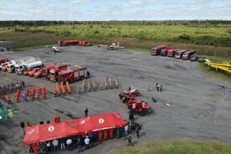 Vista aérea de treinamento de bombeiros, com veículos de emergência, tendas e equipes reunidas em uma área externa.