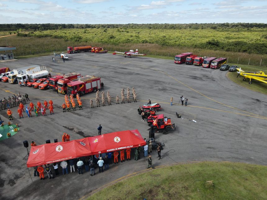 Vista aérea de treinamento de bombeiros, com veículos de emergência, tendas e equipes reunidas em uma área externa.